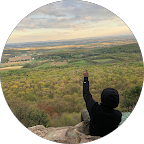 Person in black jacket and hat sitting on a rocky ledge pointing upwards, overlooking a vast green forested valley and distant fields under a cloudy sky.