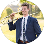 Young man in a blue suit and striped tie holding a trombone resting on his shoulder outdoors.