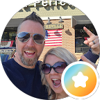 Smiling man and woman wearing sunglasses posing together outdoors in front of a building with an American flag and a sign.