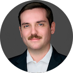 Portrait of a man with short dark hair and a mustache wearing a dark suit jacket and white shirt against a gray background.