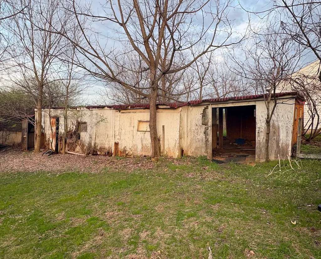 Old, weathered wooden shed with a damaged door and no leaves on surrounding trees, set on a patchy grass lawn.