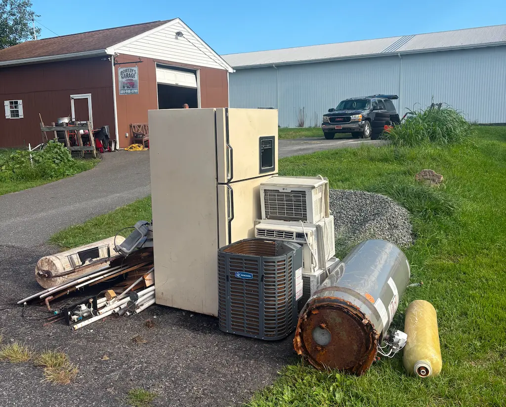 A pile of old household appliances and equipment including a beige refrigerator, several air conditioning units, and rusty cylindrical tanks piled on asphalt near a brown garage with a sign reading 'Bortz's Garage'.