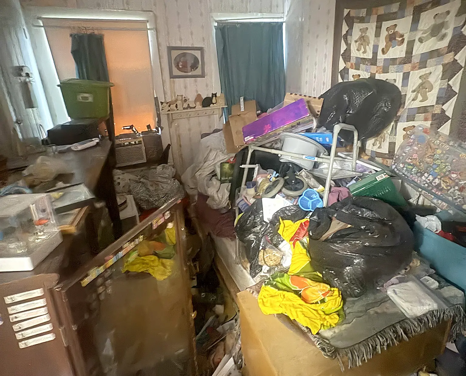 Cluttered room with a bed covered in assorted items including black trash bags, clothes, boxes, and household objects, a wooden dresser with an open glass door, and a wall hanging with teddy bear quilts.