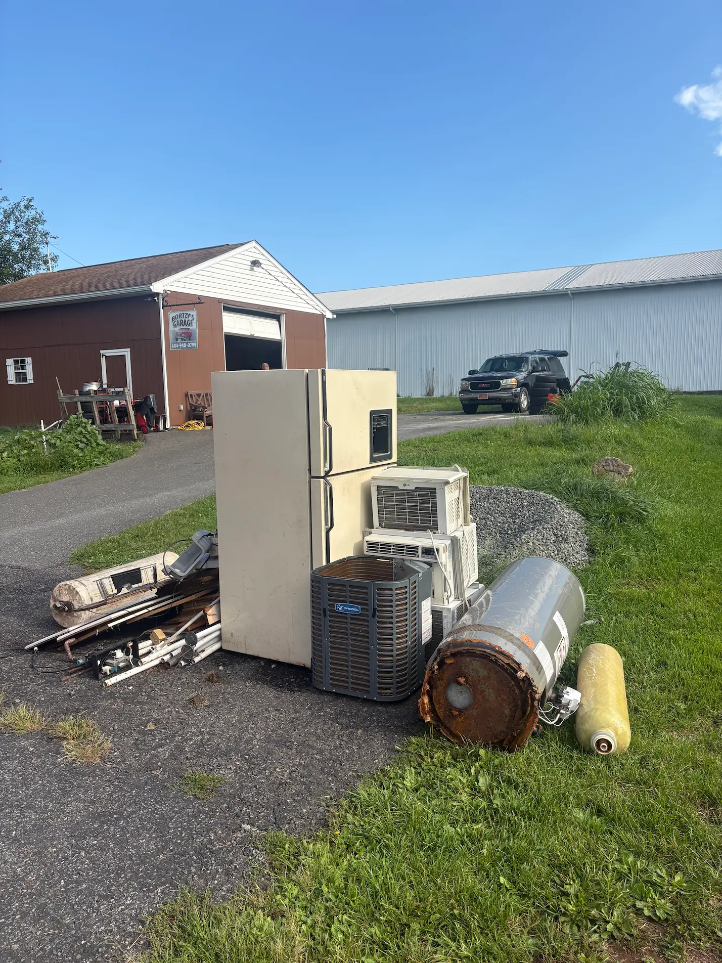 Outdoor pile of old appliances including a refrigerator, an air conditioner, a water heater, and other miscellaneous metal and plastic parts on a paved area near a brown garage and a black SUV.