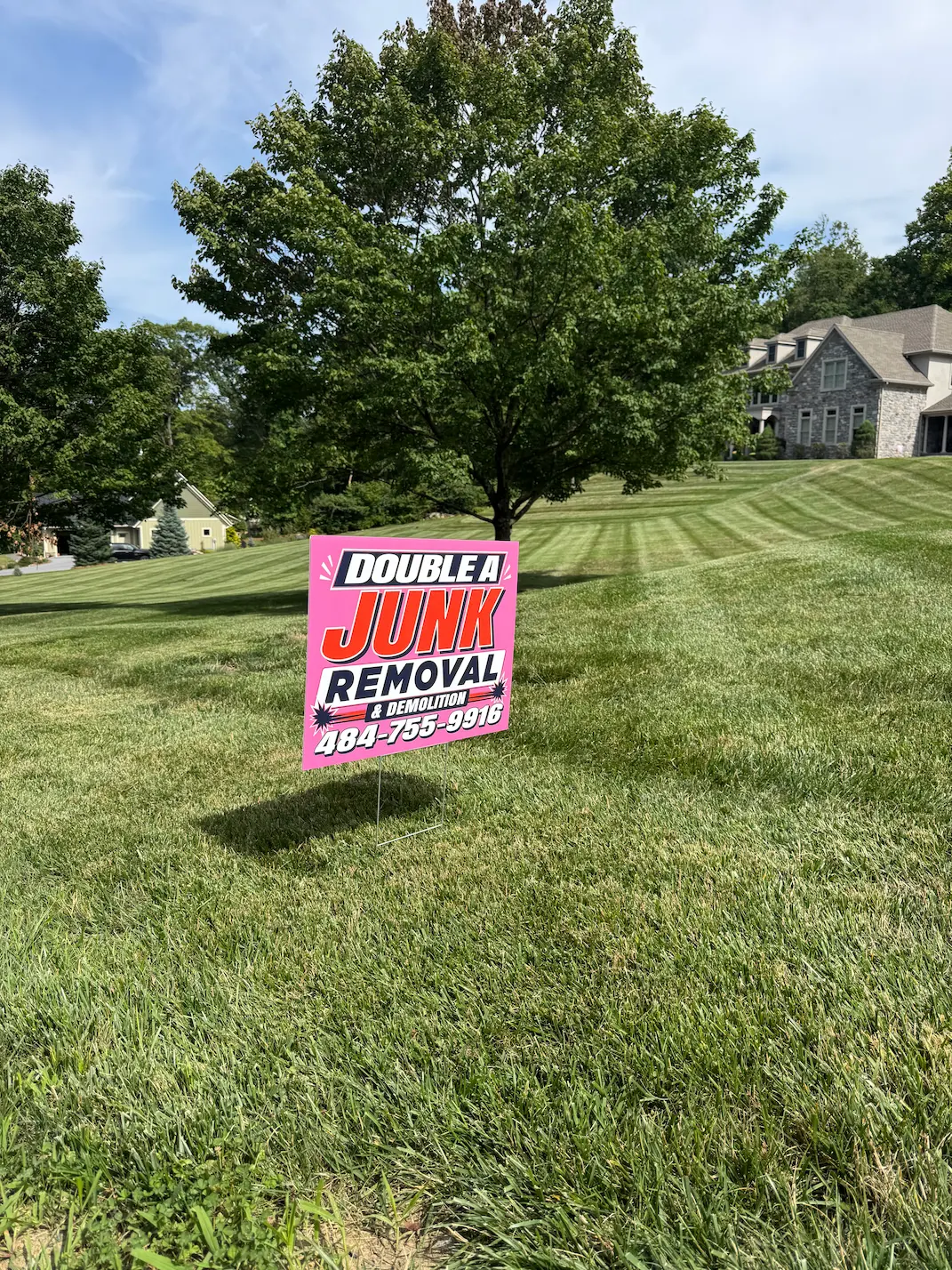 Yard sign in green grass advertising Double A Junk Removal and Demolition with phone number, set against a backdrop of trees and residential houses under blue sky.