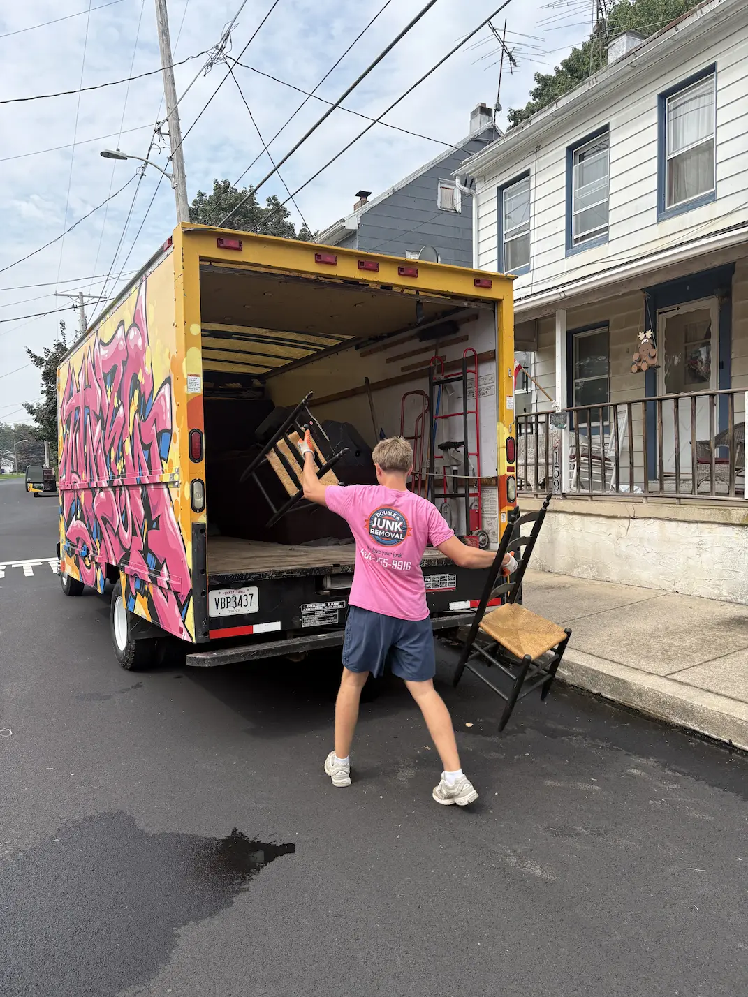 Person wearing a pink 'Double A Junk Removal' shirt loading two wooden chairs into the back of a colorful yellow and pink junk removal truck parked on a residential street.