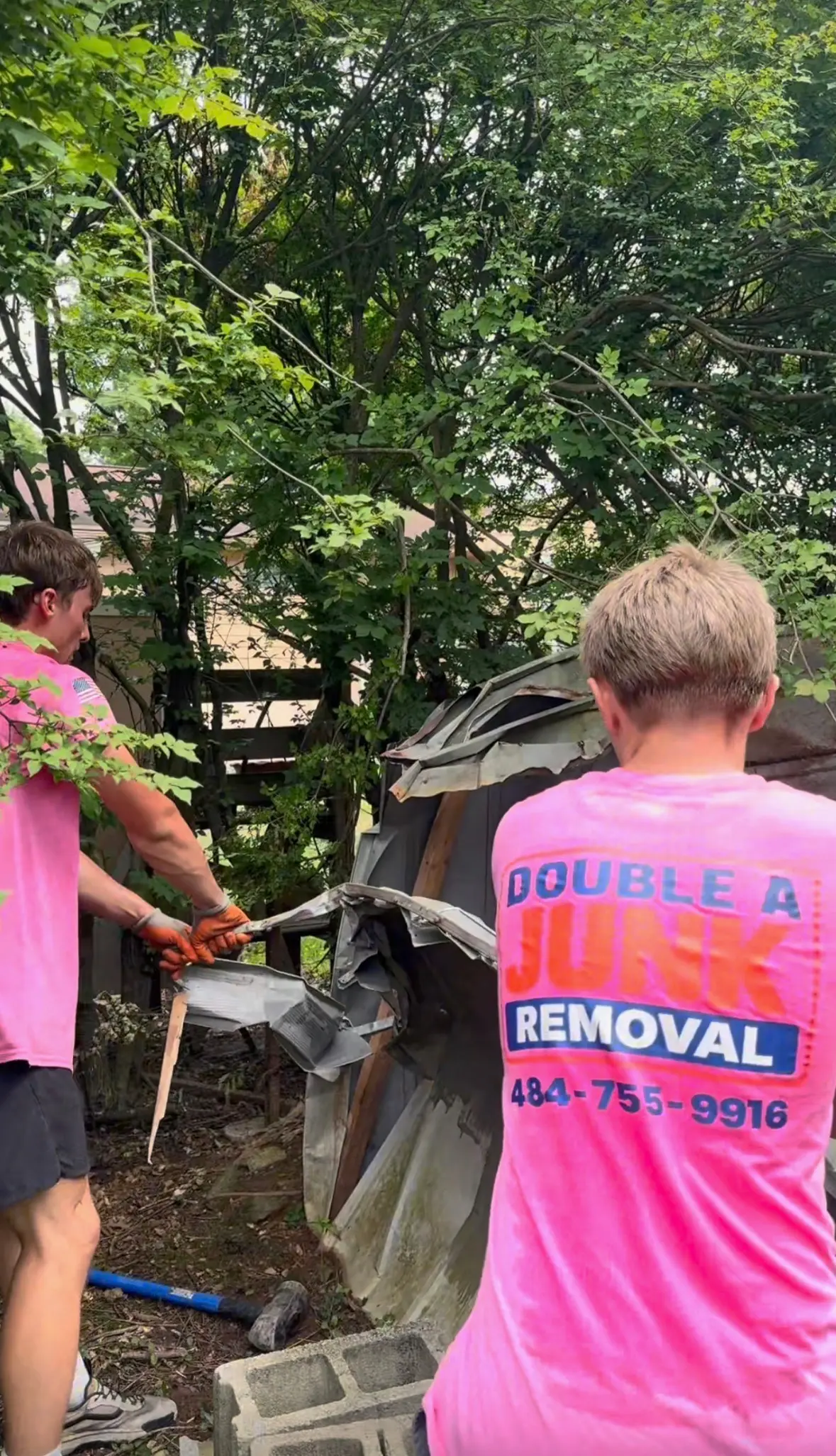Two workers in bright pink shirts demolition a shed outdoors, surrounded by trees and cinder blocks, with one worker pulling metal off the structure and a sledgehammer on the ground.