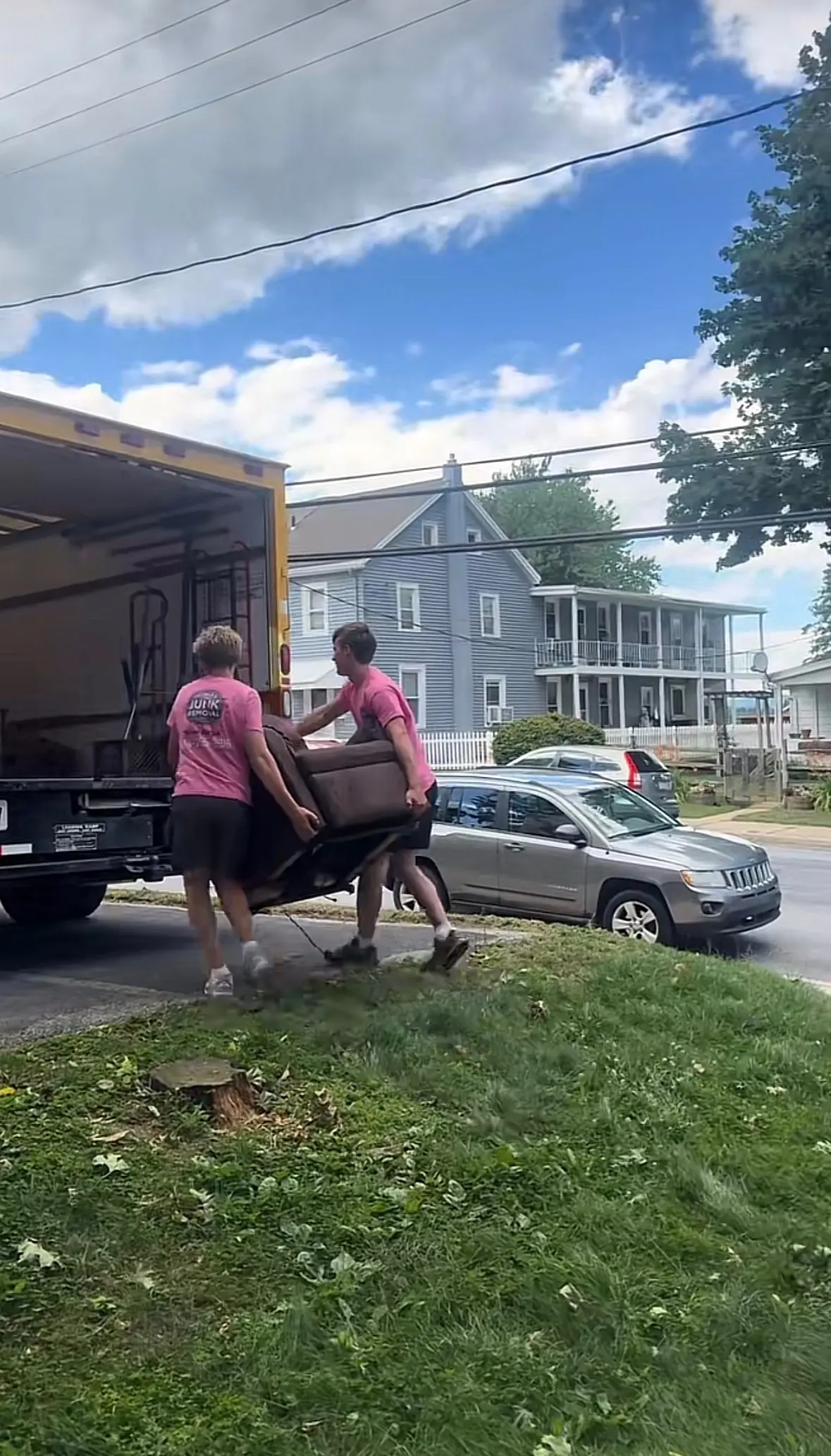 Two men wearing matching pink shirts load a brown chair into the back of a yellow moving truck on a residential street with houses and cars visible.