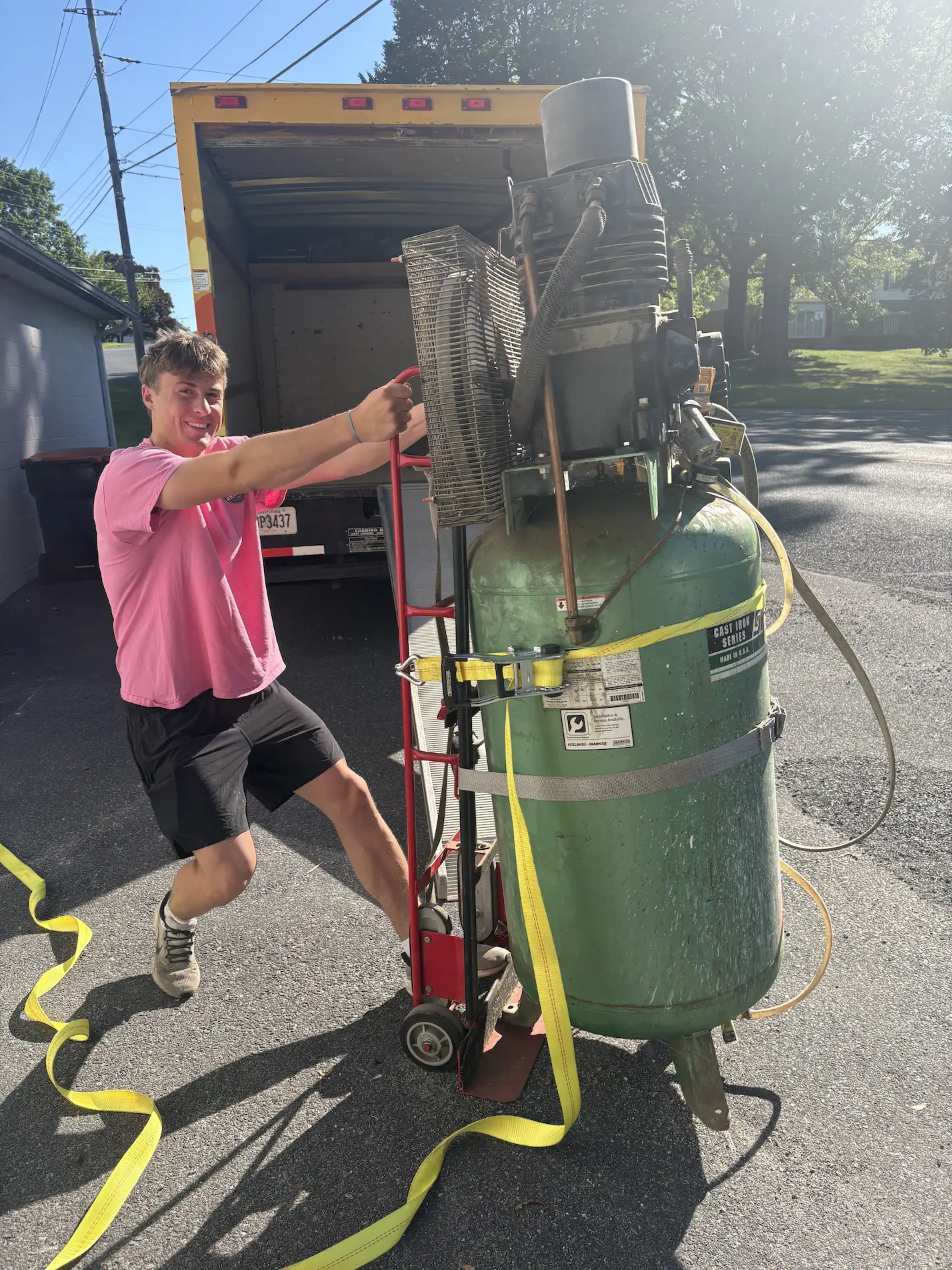 A young man in a pink shirt and black shorts smiling while moving a large green industrial tank secured with straps on a red hand truck near an open yellow truck.