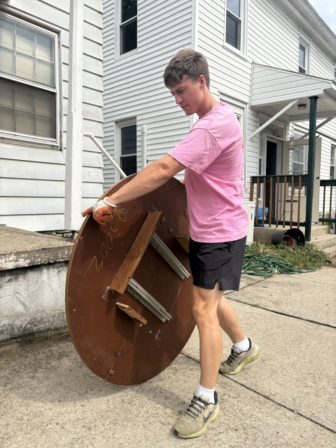 Young man in a pink shirt and black shorts lifts a round wooden table upside down outside a white house on a concrete sidewalk.