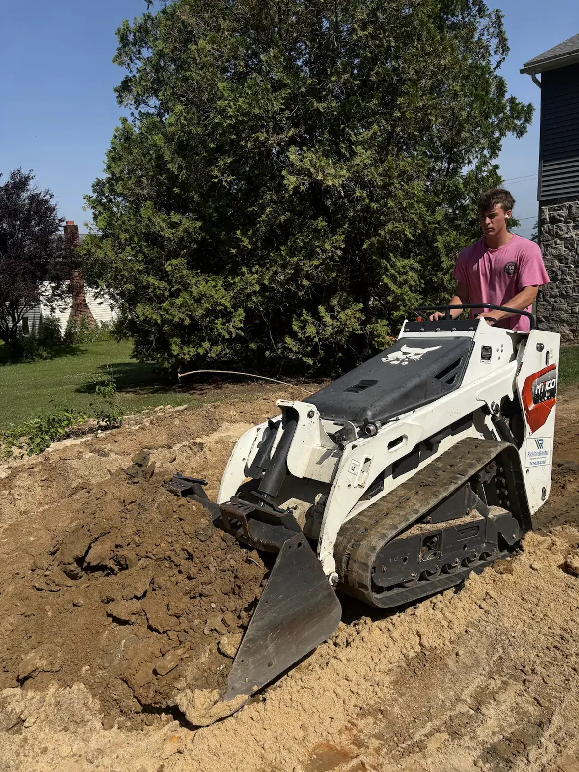Young man operating a compact Bobcat MT100 mini track loader to move a pile of dirt in a yard with green trees and grass in the background.