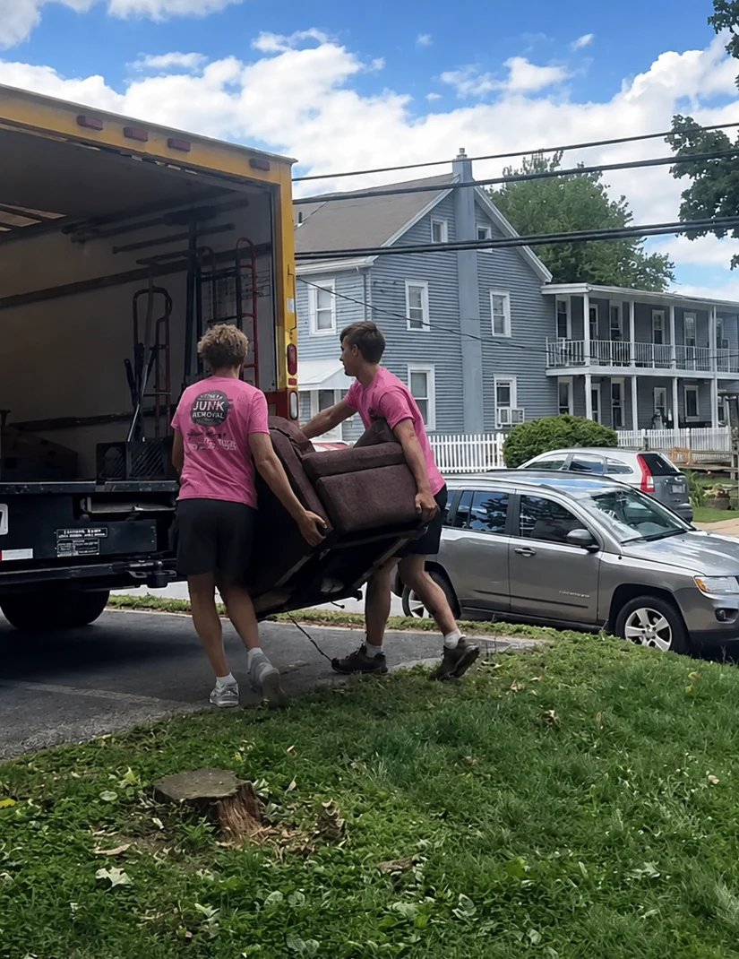 Two men wearing pink shirts carrying a brown couch into the back of a yellow moving truck on a residential street with houses and parked cars in the background.