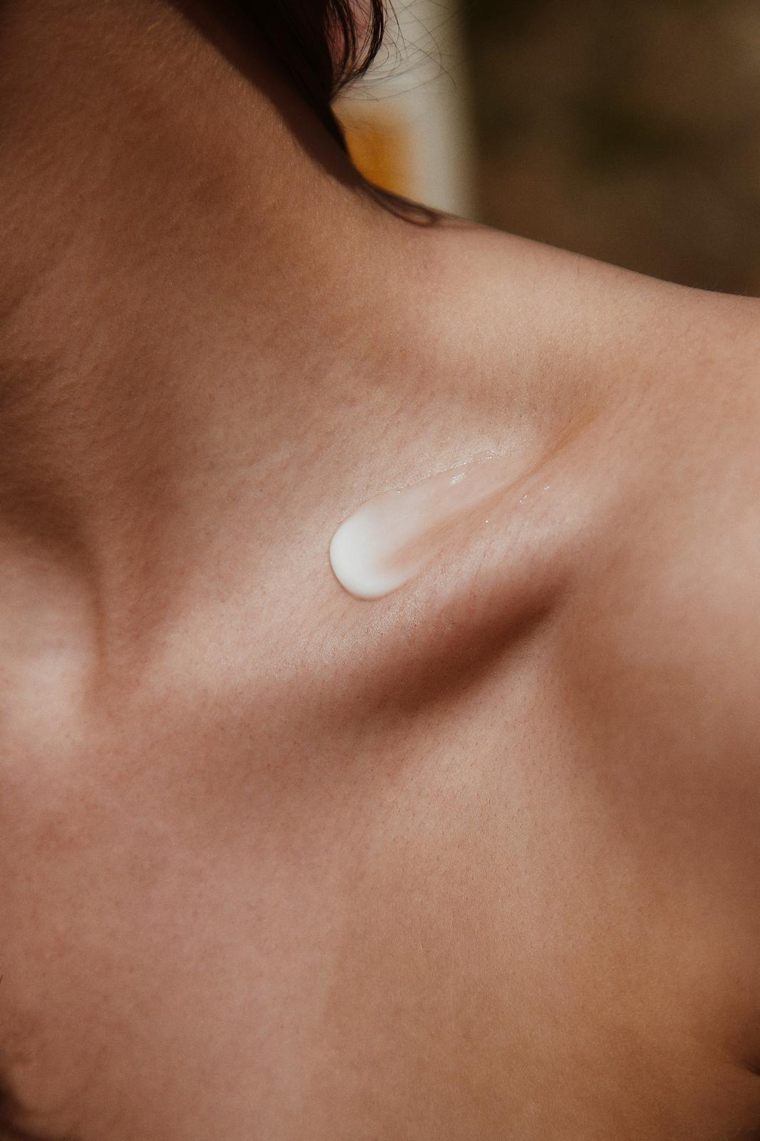 Close-up of a person's shoulder and upper chest with a small dollop of white lotion on the skin near the collarbone.