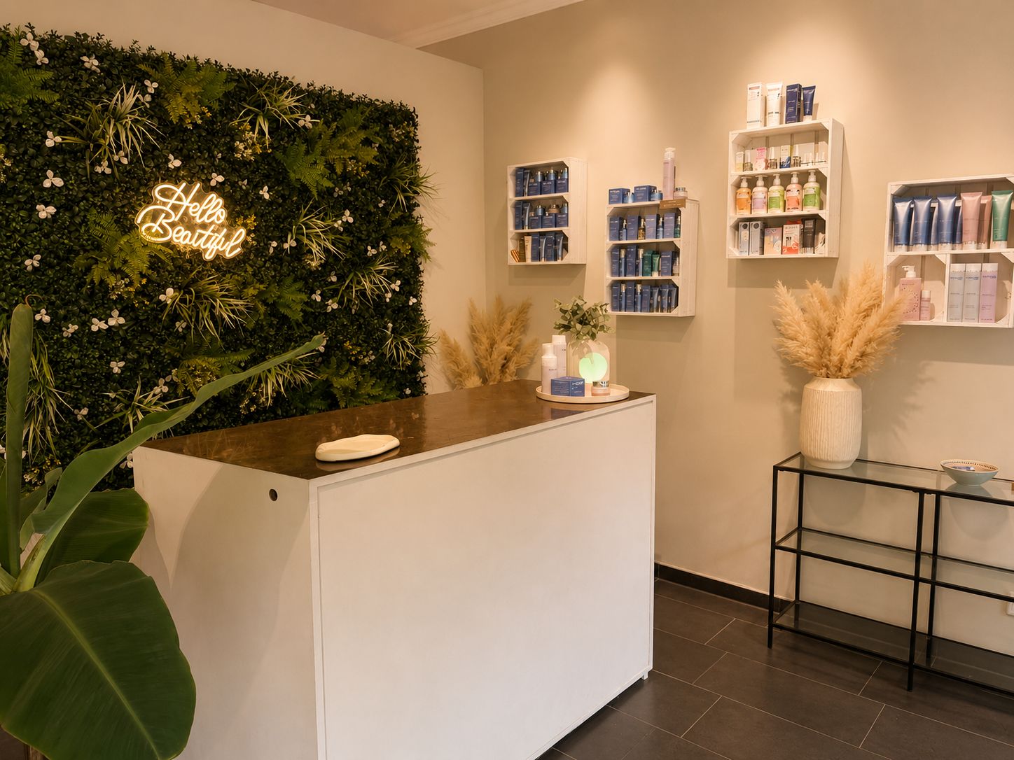 A modern beauty studio reception area with a white desk topped with a dark surface, a green plant in the foreground, and a lush green wall behind the desk with a glowing neon sign reading 'Hello Beautiful.' Several shelves on the wall display assorted skincare products in colorful packaging, and beige pampas grass in vases add a natural decorative touch.