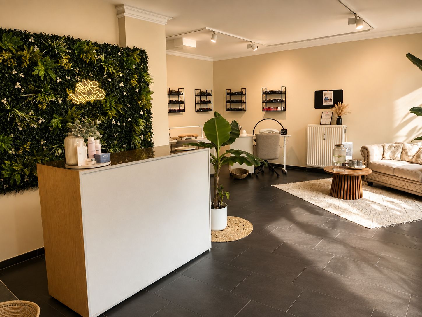 Bright, modern beauty salon interior featuring a white reception desk with skincare products, a green plant on a round woven mat, a green foliage wall with a neon sign, multiple nail polish racks on the back wall, two manicure stations with chairs, and a cozy seating area with a beige couch, wooden round table on a cream rug, and a beverage dispenser with glasses.