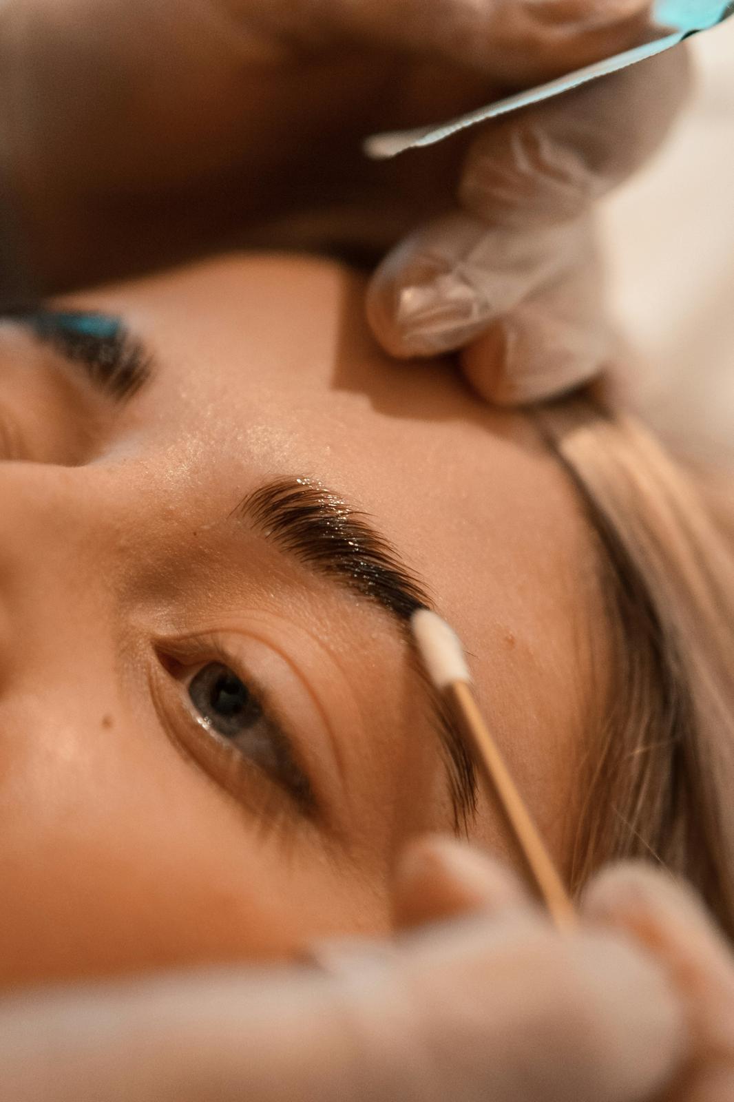 Close-up of a person's face undergoing an eyebrow grooming treatment with a gloved hand using a cotton swab and a tool near the eyebrow.