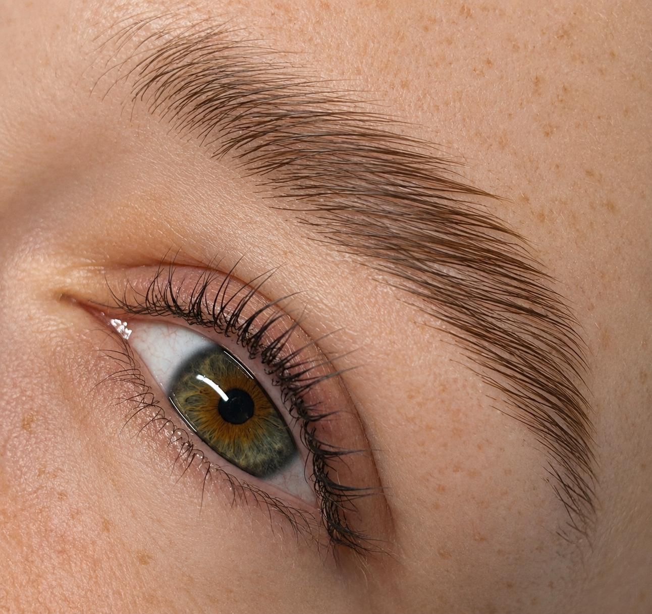 Close-up of a green eye with brown and gold flecks surrounded by natural lashes and well-groomed brown eyebrow on light freckled skin.