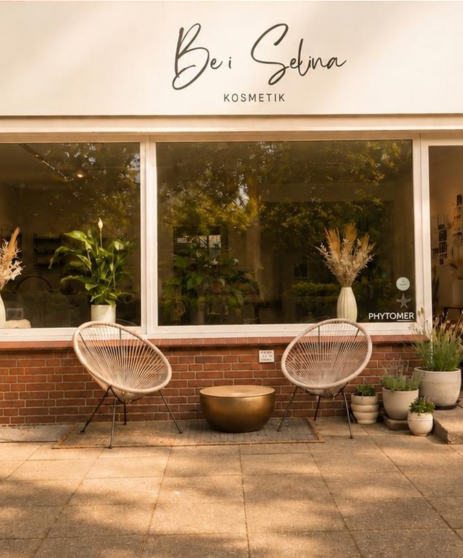Exterior of a cosmetic store named 'Bei Selina Kosmetik' with a large window showing green plants inside, two modern outdoor chairs, a brass round table, and several potted plants arranged in front on a tiled sidewalk.