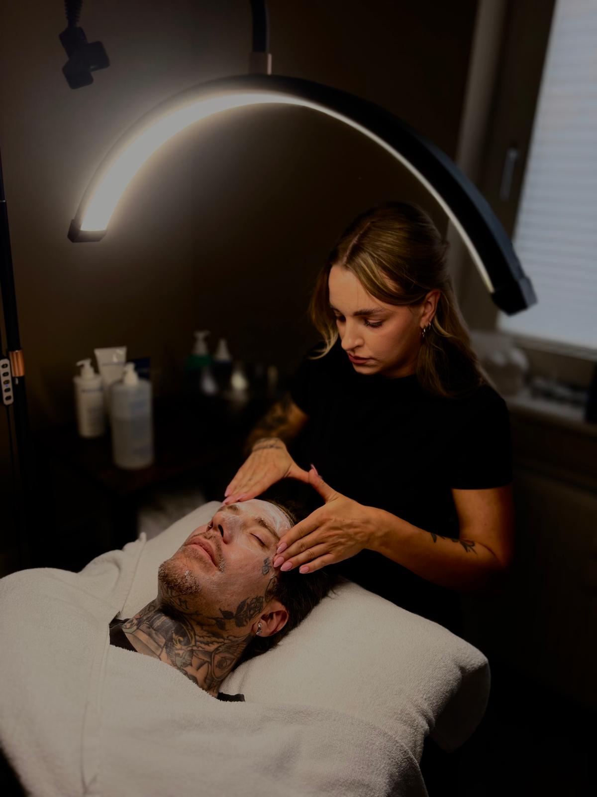 A tattooed man lies relaxed with eyes closed on a spa bed covered with a white towel as a woman carefully massages cream on his face under an overhead curved light in a softly lit room.