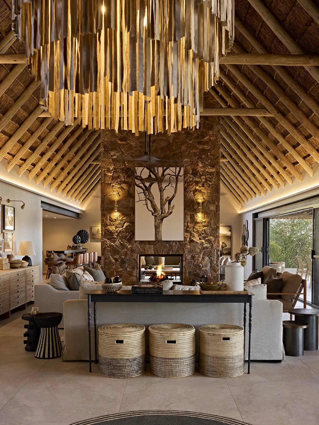 Hospitality interior design showcasing a Game lodge lounge with a sculptural timber chandelier, stone fireplace and woven basket seating beneath a thatched roof.