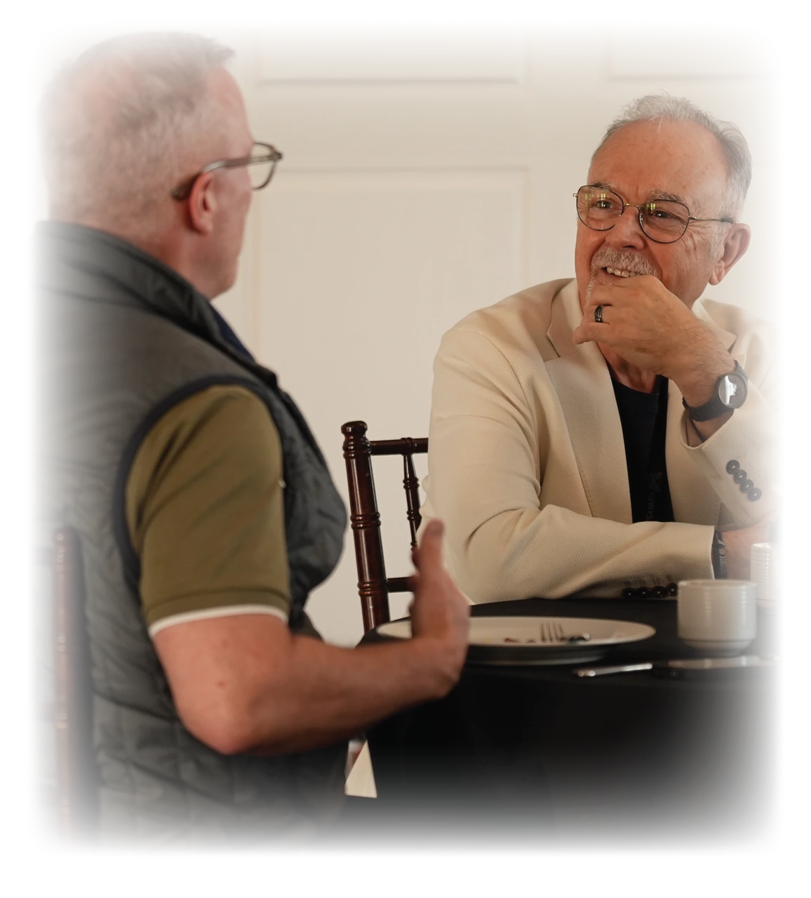 Two men in conversation across a table, one listening with his hand at his chin