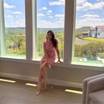 Woman in a pink dress sitting on a wide white windowsill with three tall windows showing a bright outdoor view with green trees, blue sky, and houses.