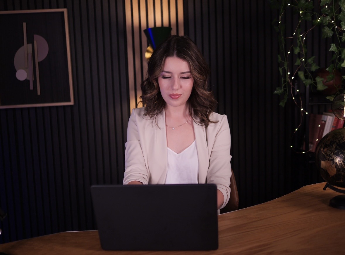 A woman with brown wavy hair wearing a white blazer sits at a wooden desk, working on a black laptop, in a dimly lit room with dark vertical panels, a framed abstract artwork, a globe, and plants with decorative string lights.