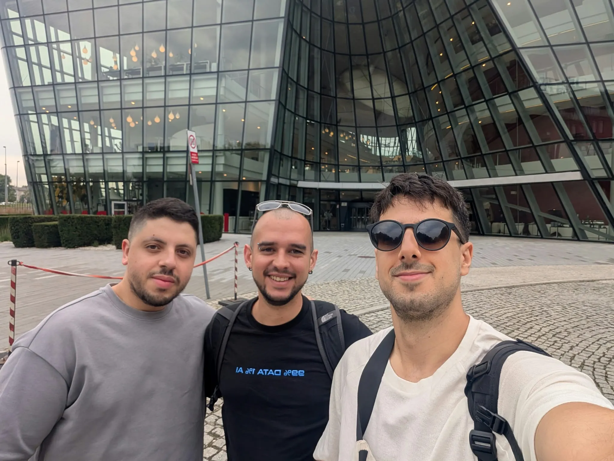 Three young men smiling and posing for a selfie in front of a modern glass building with reflective windows and angular architecture. Two men wear backpacks and one wears sunglasses.