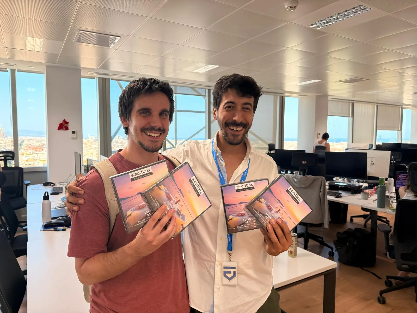 Two men smiling in an office setting, each holding three Smartbox gift boxes featuring a sunset luxury stay theme, with computer desks and large windows in the background showing a city view.