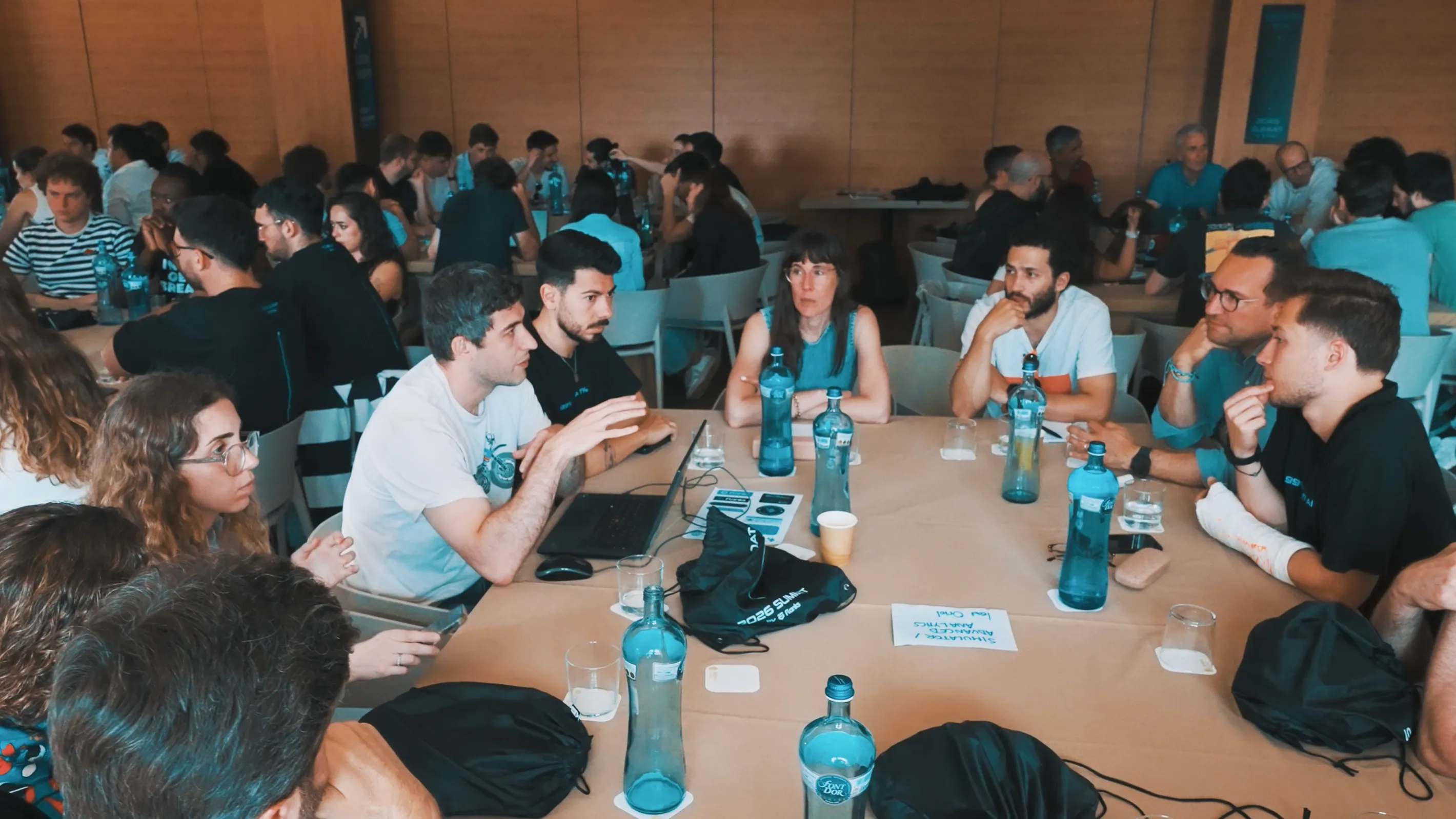 Group of people seated around tables in a discussion or meeting setting, with water bottles, papers, and laptop visible, in a room with wooden panel walls.