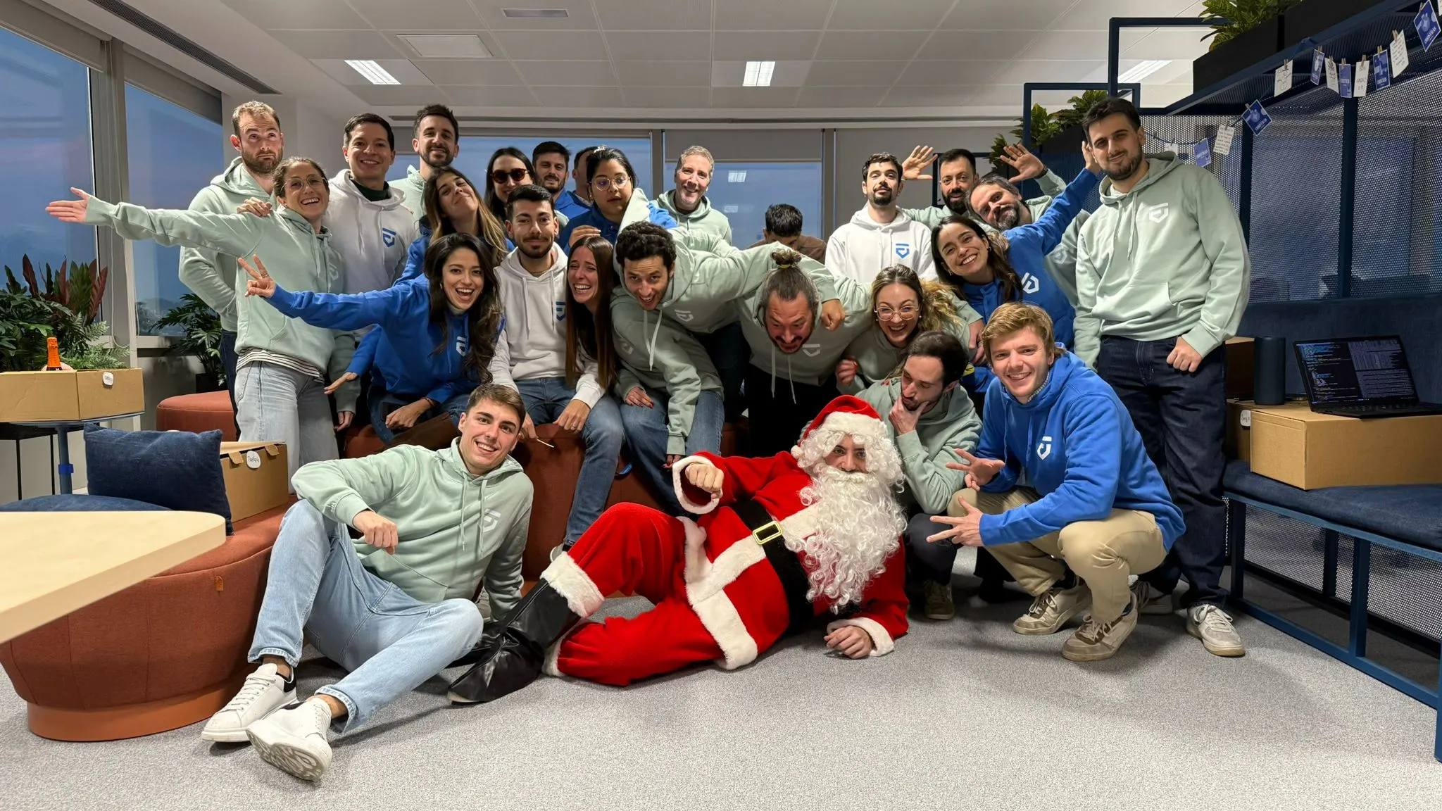 Group photo of young adults in an office setting posing cheerfully around a person dressed as Santa Claus lying on the floor in front of them. Most people wear green or blue hoodies with a white logo, smiling and making playful gestures.