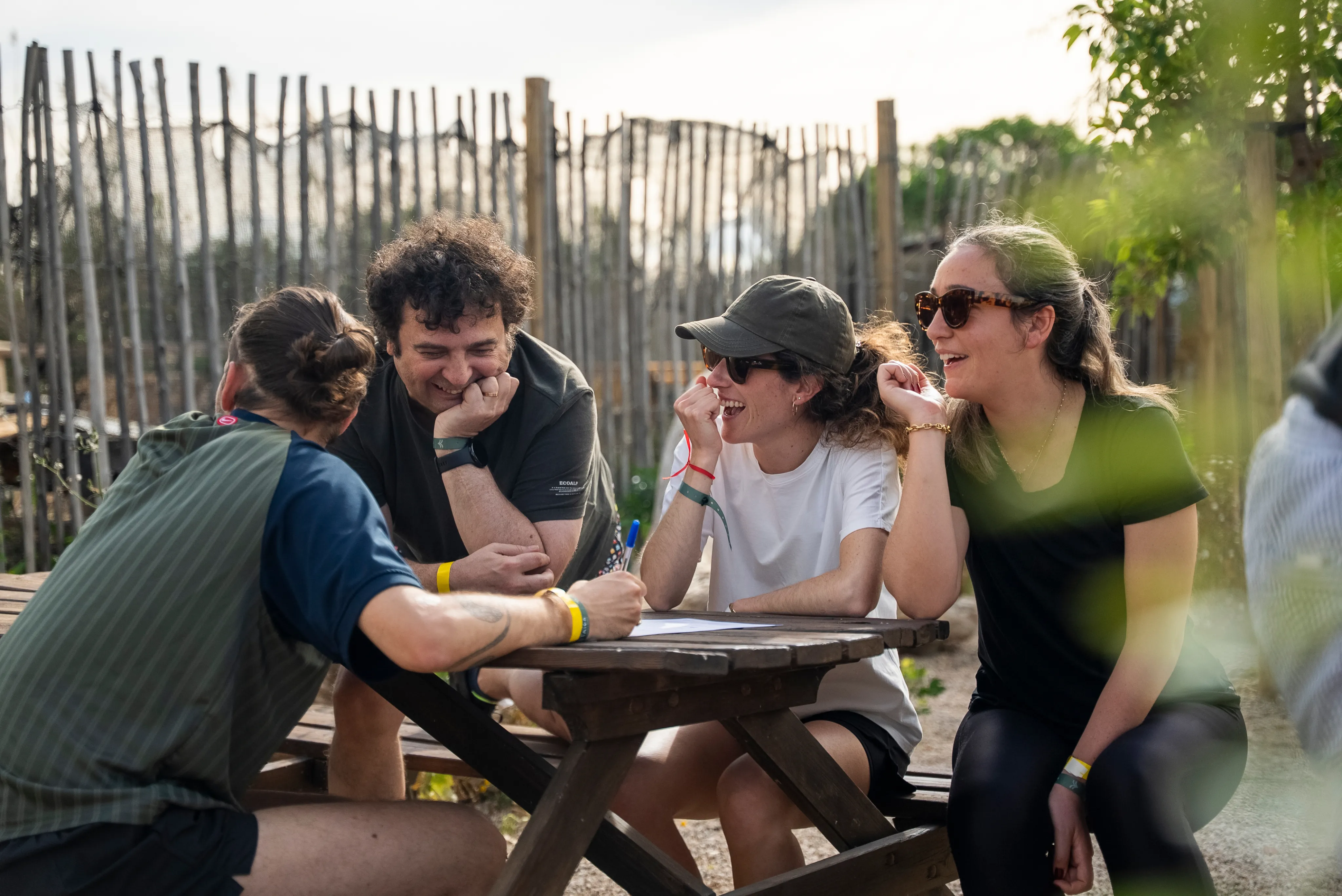 Four people sitting at a wooden picnic table outdoors, engaging in a lively conversation and smiling, with one person holding a pen and writing on paper.