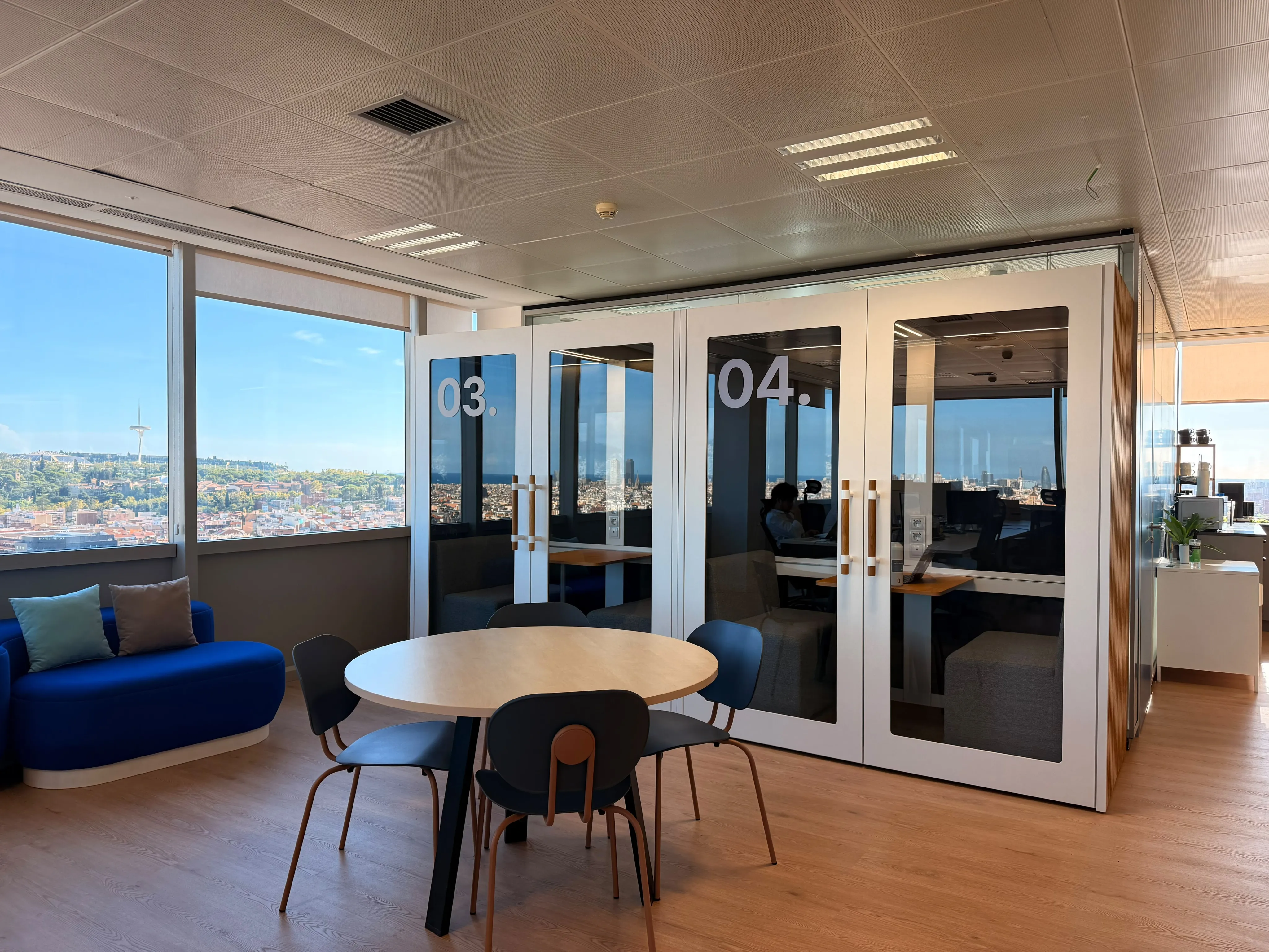 Modern office space with two phone booths labeled 03 and 04, a round wooden table surrounded by four black chairs, and a blue sofa by large windows showing a cityscape with a tower and greenery.