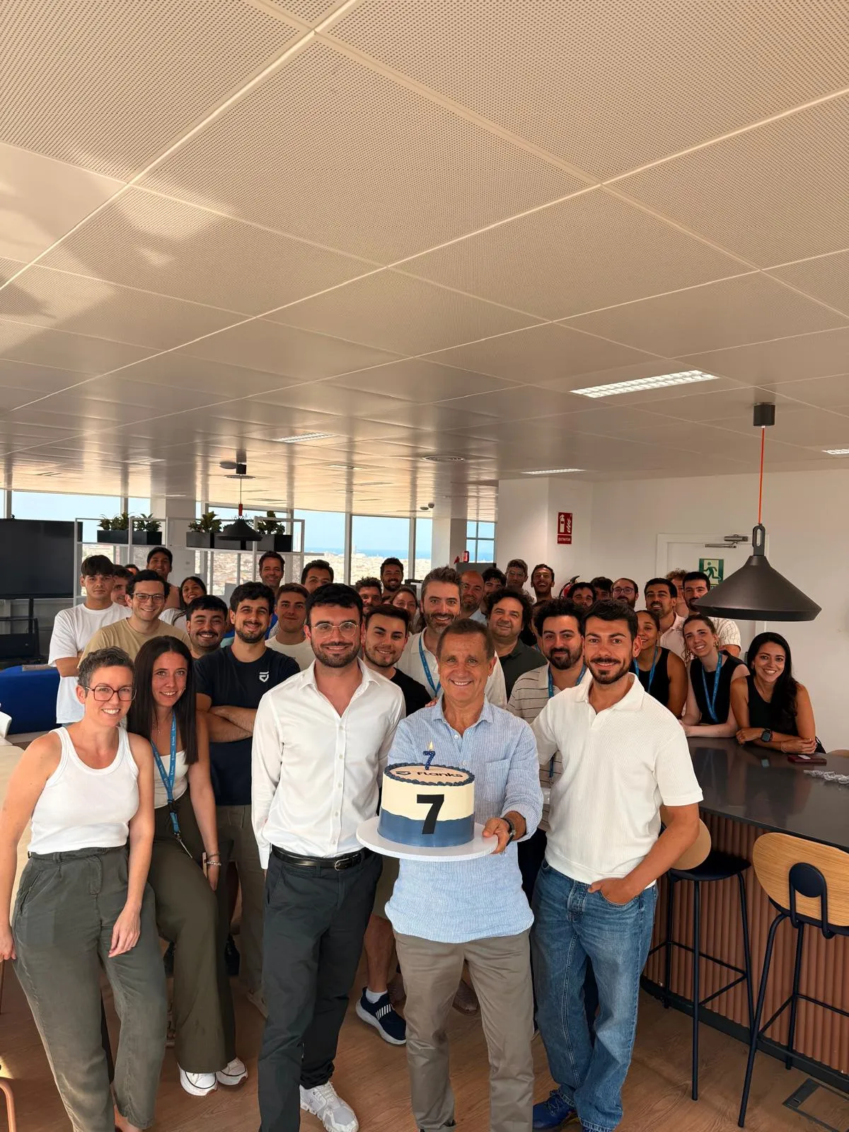 Group of diverse office colleagues gathered in a modern workspace, smiling towards the camera as an older man in front holds a cake with the number 7 on it, celebrating an anniversary or milestone.