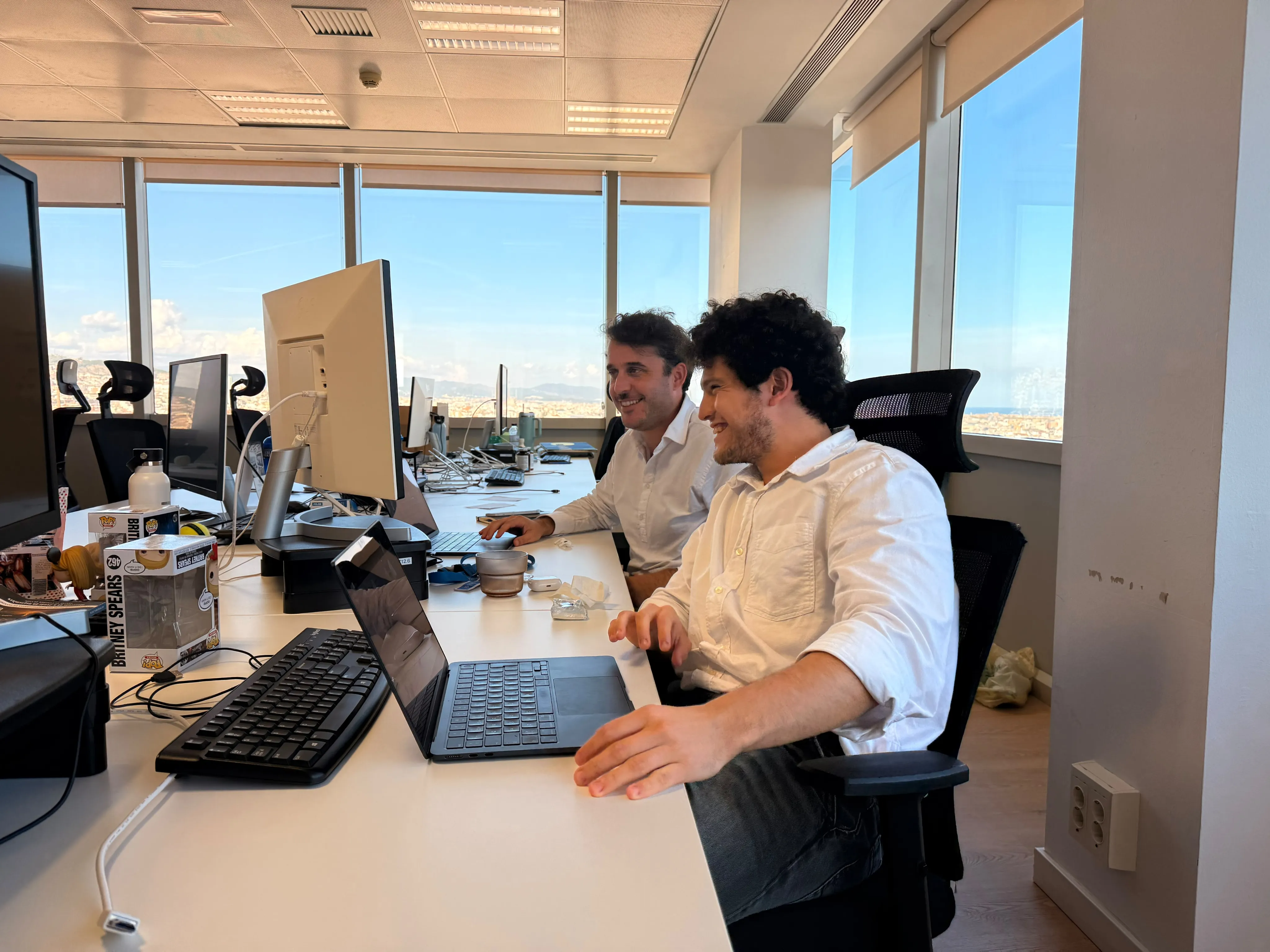 Two men in white shirts sitting at a long office desk with multiple monitors and laptops, smiling and looking at a laptop screen, with large windows showing a cityscape in the background.
