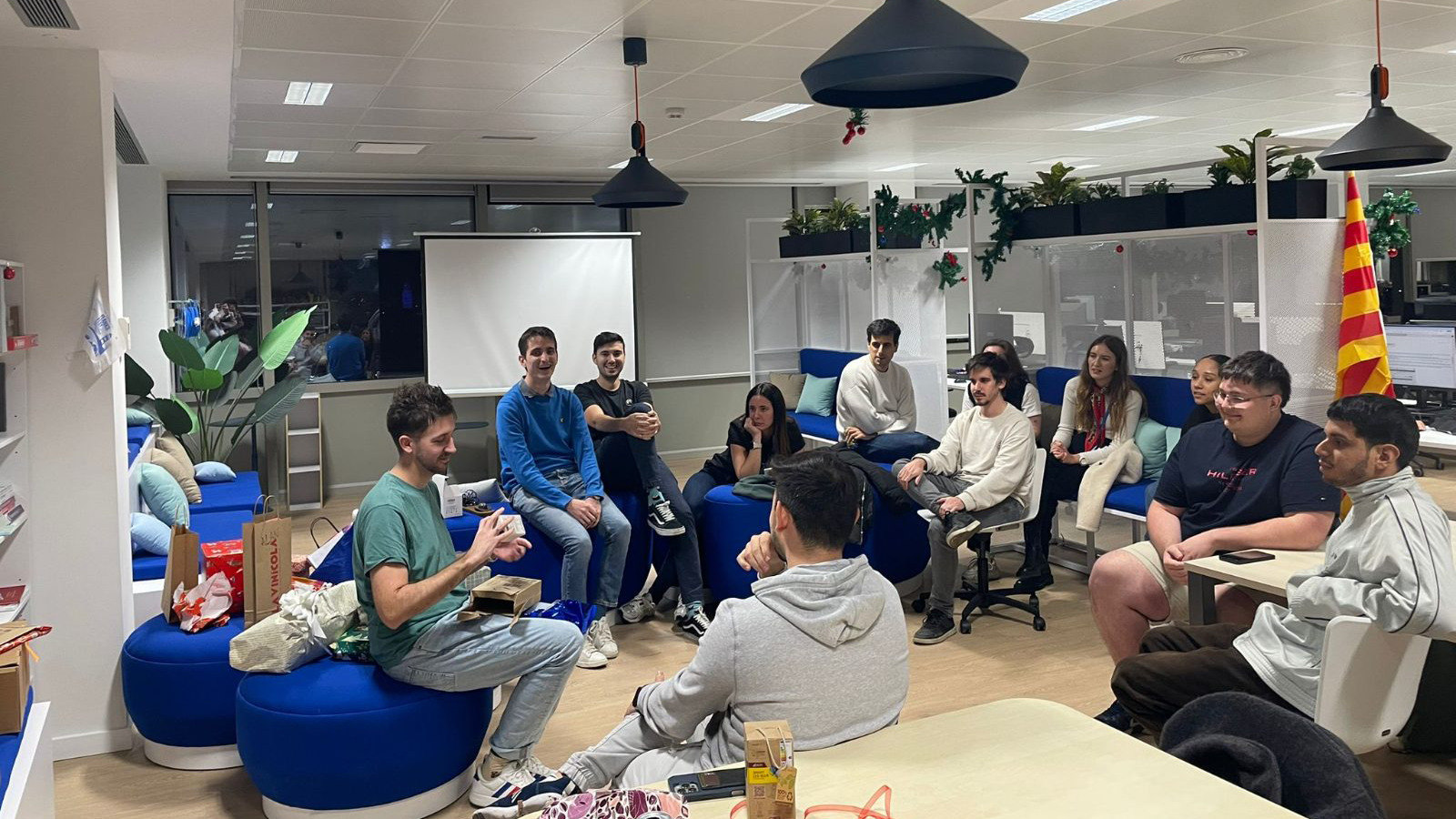 A group of people sitting in a modern office space engaged in conversation or a casual meeting, with blue cushioned seats, plants, a projector screen, and a yellow and red striped flag in the background.