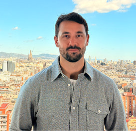 Man with dark hair and beard wearing a gray button-up shirt standing in front of a cityscape with buildings and mountains under a blue sky with some clouds.