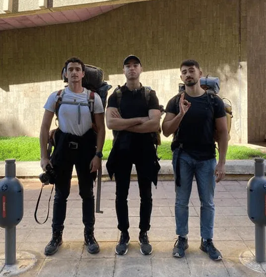 Trois jeunes hommes debout, portant des sacs à dos de randonnée, devant un mur beige avec des zones d'ombre et de lumière.