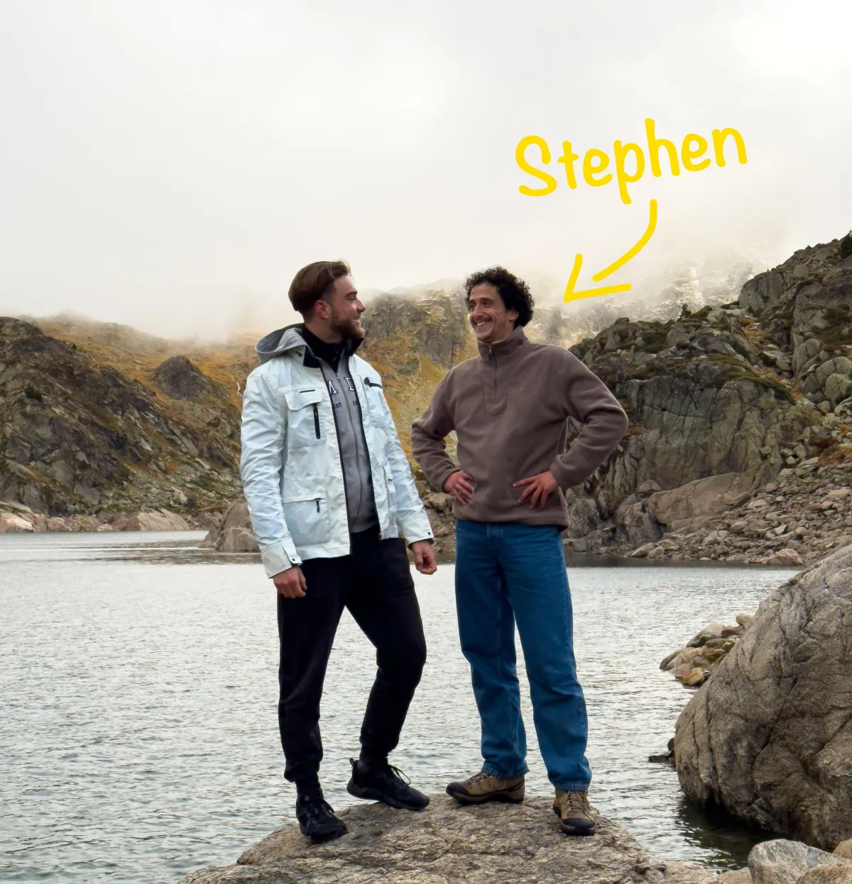 Deux hommes souriants debout sur un rocher près d'un lac de montagne, entourés de rochers et de collines brumeuses, avec une annotation jaune indiquant "Stephen" pour l'homme à droite.
