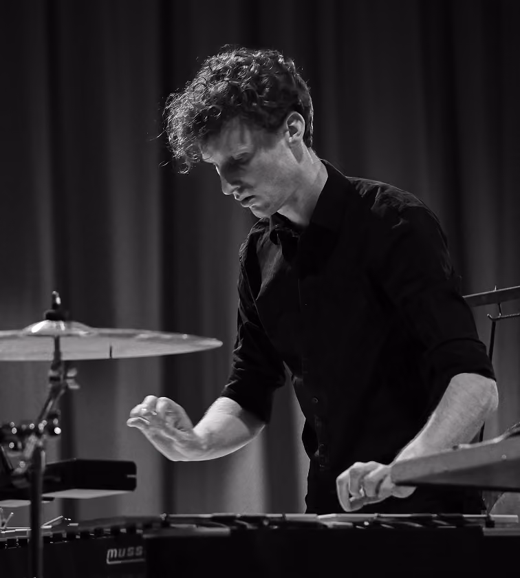 Un musicien aux cheveux bouclés joue du vibraphone avec une main levée et l'autre frappant les touches, devant un rideau et une cymbale en arrière-plan.