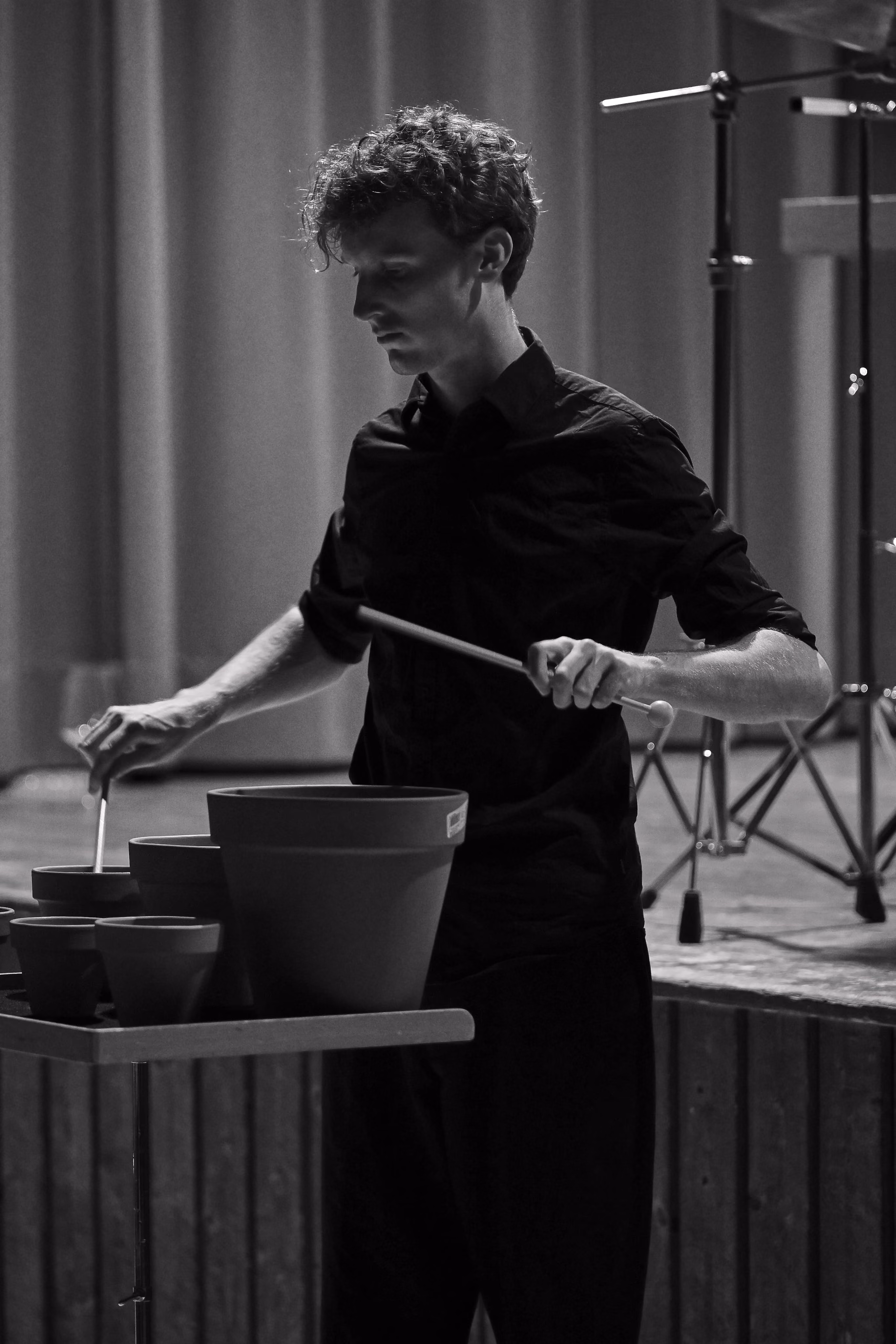 Un homme aux cheveux bouclés en chemise noire joue des pots de fleurs comme des instruments de musique sur une scène.