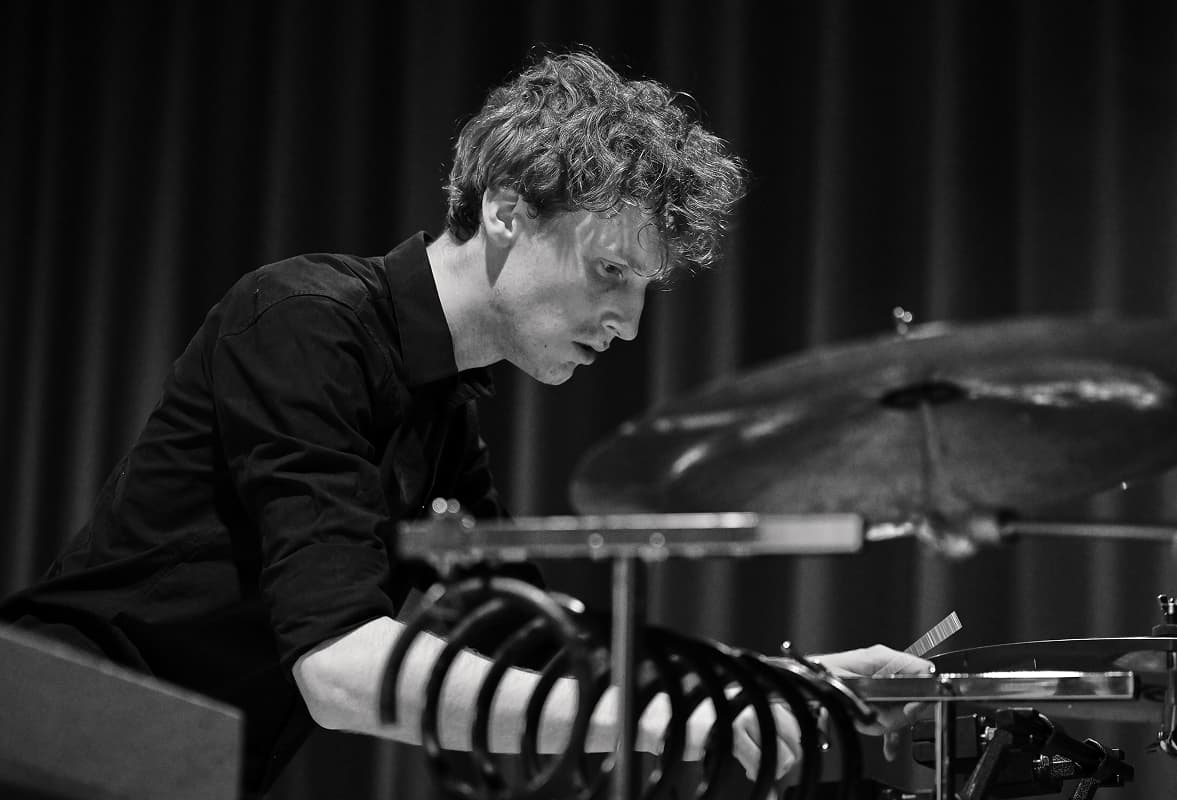 Homme jouant de la batterie lors d'une performance, concentré sur son instrument, en chemise noire, avec des rideaux sombres à l'arrière-plan.