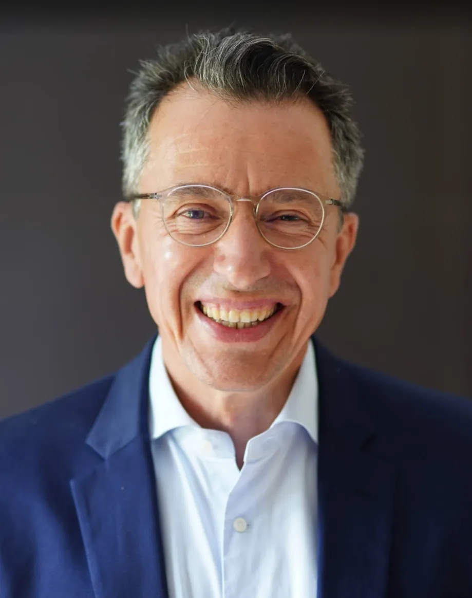 Middle-aged man with gray hair and glasses smiling broadly, wearing a dark blue blazer over a white shirt, with a dark blurred background.