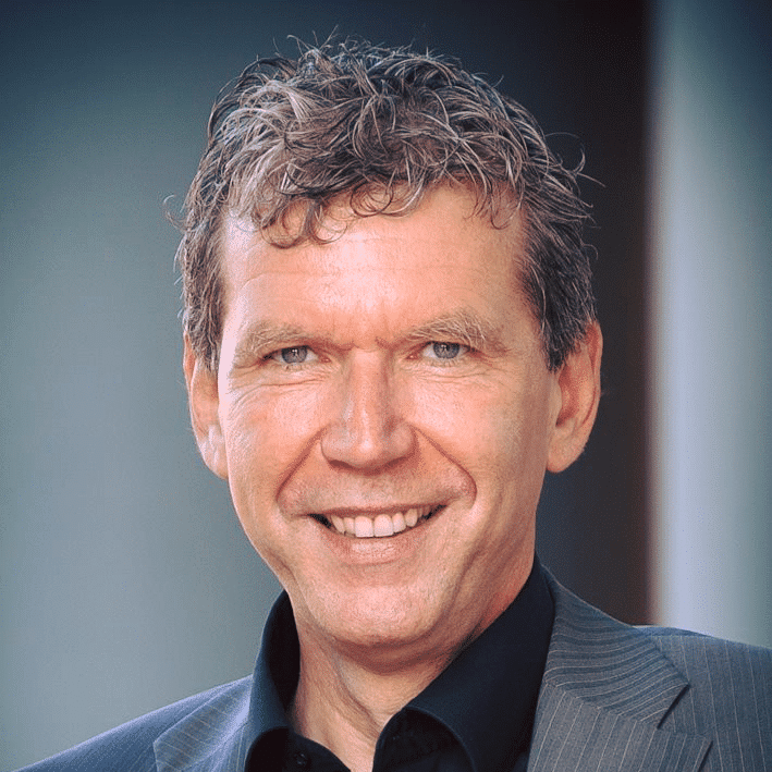 Man with short curly brown hair smiling, wearing a dark shirt and gray suit jacket against a blurred background.