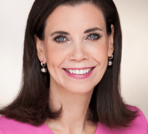 Portrait of a smiling woman with dark hair, wearing pearl earrings and a pink top, against a light background.