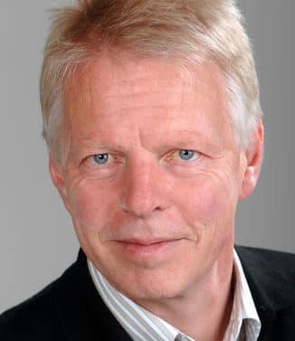 Portrait of a middle-aged man with light blonde hair, blue eyes, and fair skin wearing a black jacket and striped shirt, looking directly at the camera with a neutral expression.