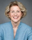 Smiling middle-aged woman with short curly blonde hair, wearing glasses and a light blue blazer, against a gray background.