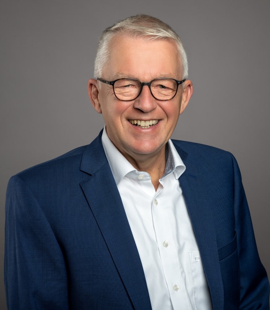 Senior man with short white hair and glasses smiling, wearing a white shirt and blue blazer, against a gray background.