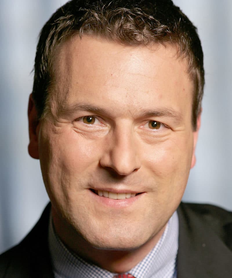 Close-up portrait of a smiling middle-aged man with short curly brown hair and light skin wearing a checkered shirt, red tie, and dark jacket.
