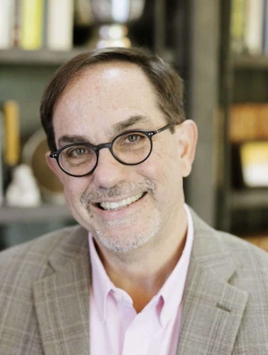 Smiling middle-aged man wearing round glasses, a light pink shirt, and a gray plaid blazer, with a blurred background of shelves and books.